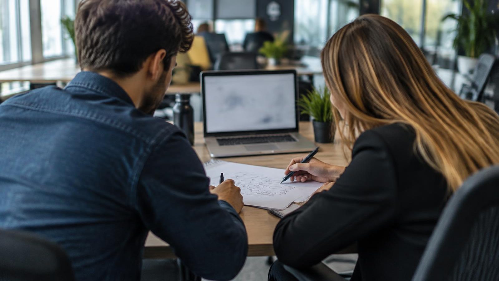 A software developer and operations manager sketch a workflow on paper beside a laptop in a modern daylight office